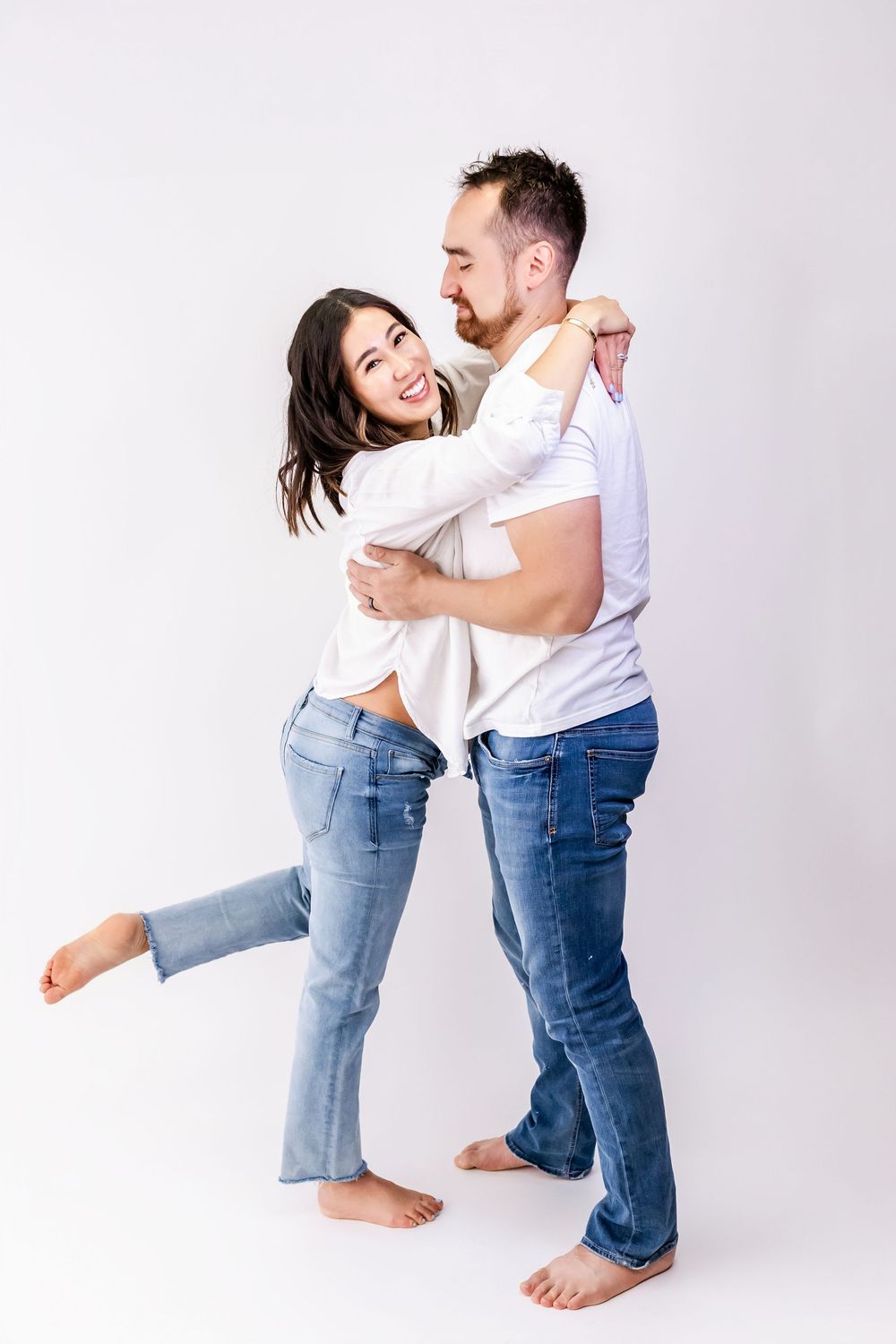 A couple wearing white shirts and blue jeans embrace joyfully against a white backdrop.