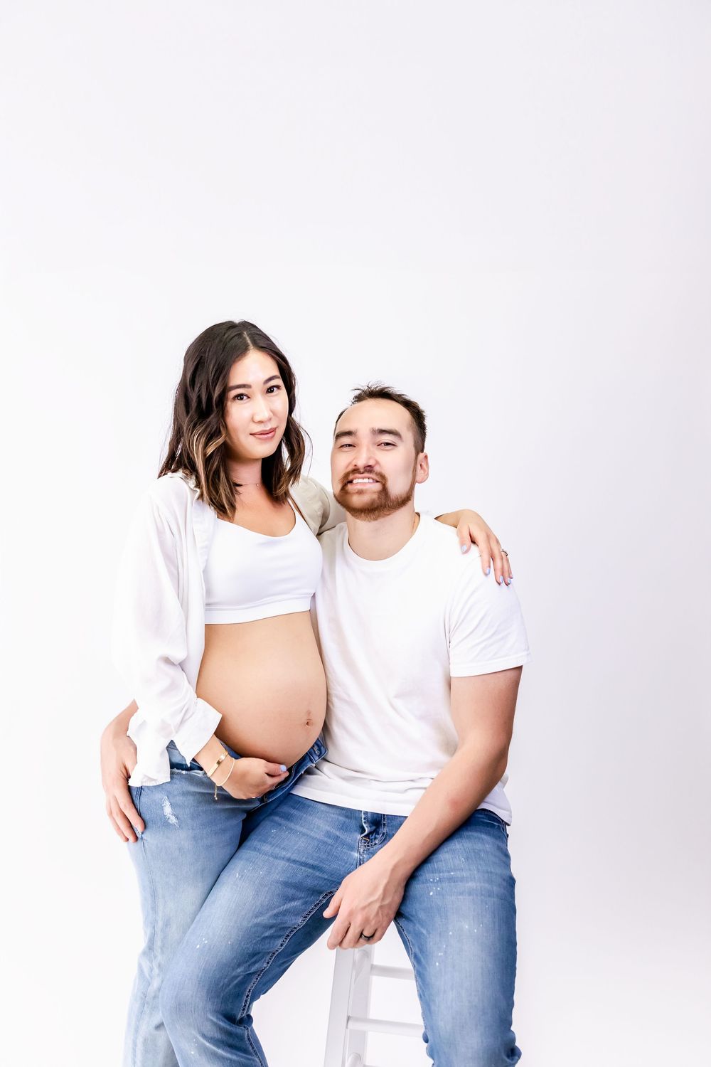 Expecting couple in white shirts and jeans pose together for maternity photo against white background.