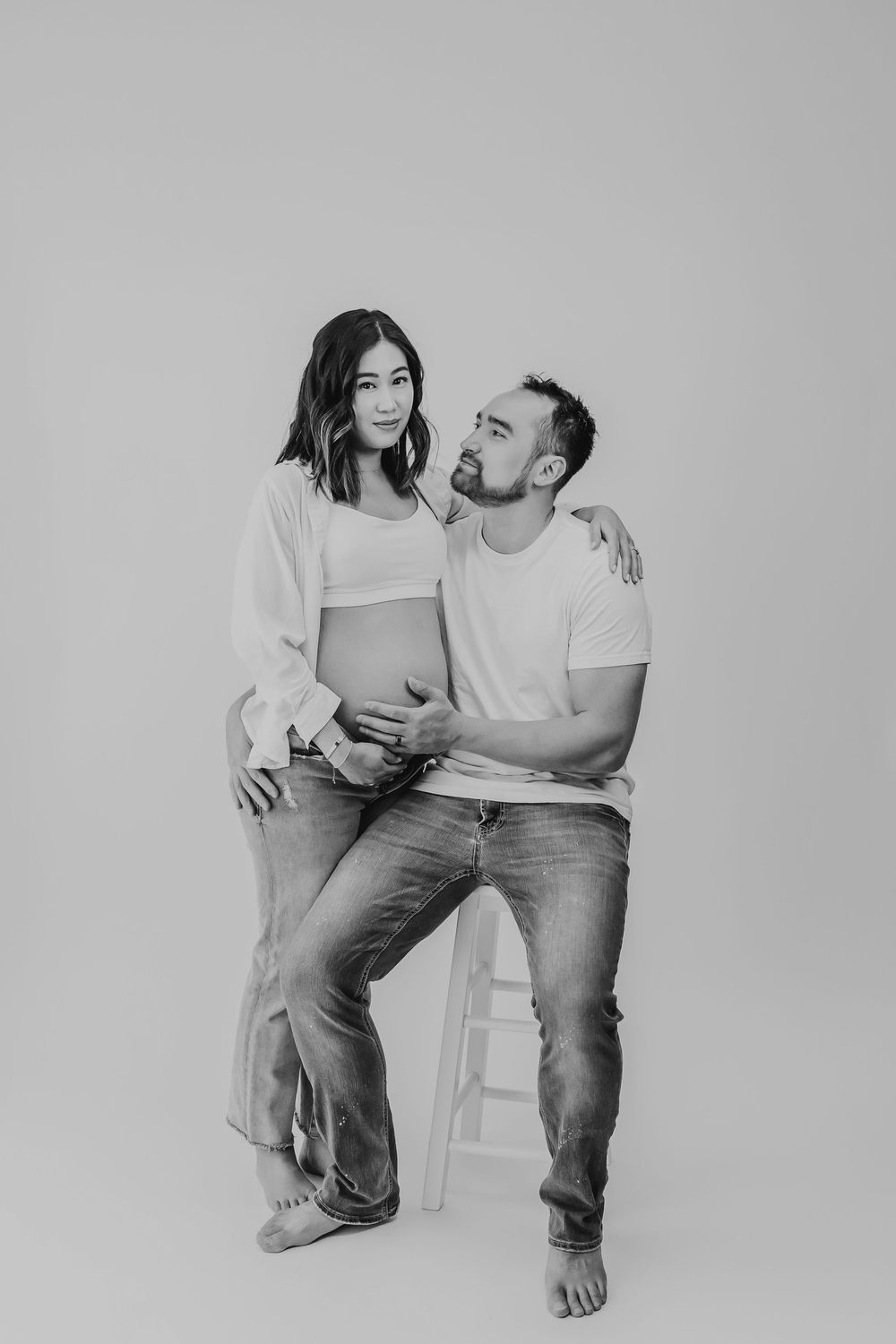 A couple shares an intimate monochrome moment while sitting on a stool against a gray background.