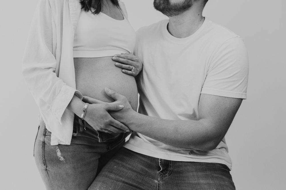 Black and white intimate maternity photo of couple in casual clothing showing affection and touching pregnant belly.