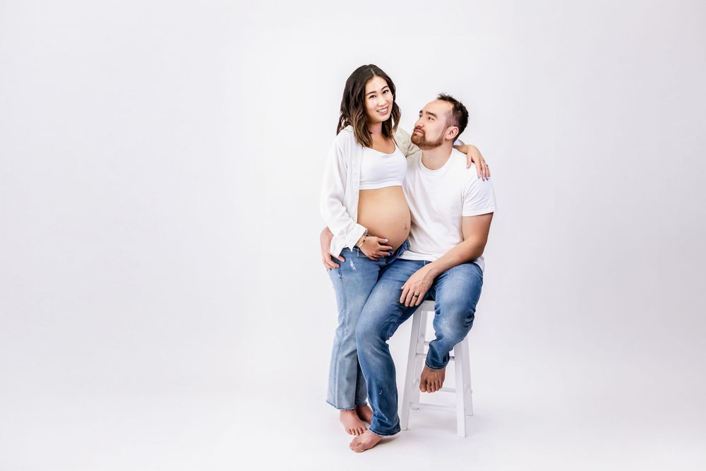 A pregnant woman wearing white shows her baby bump while sitting with a partner on a white chair against plain backdrop.