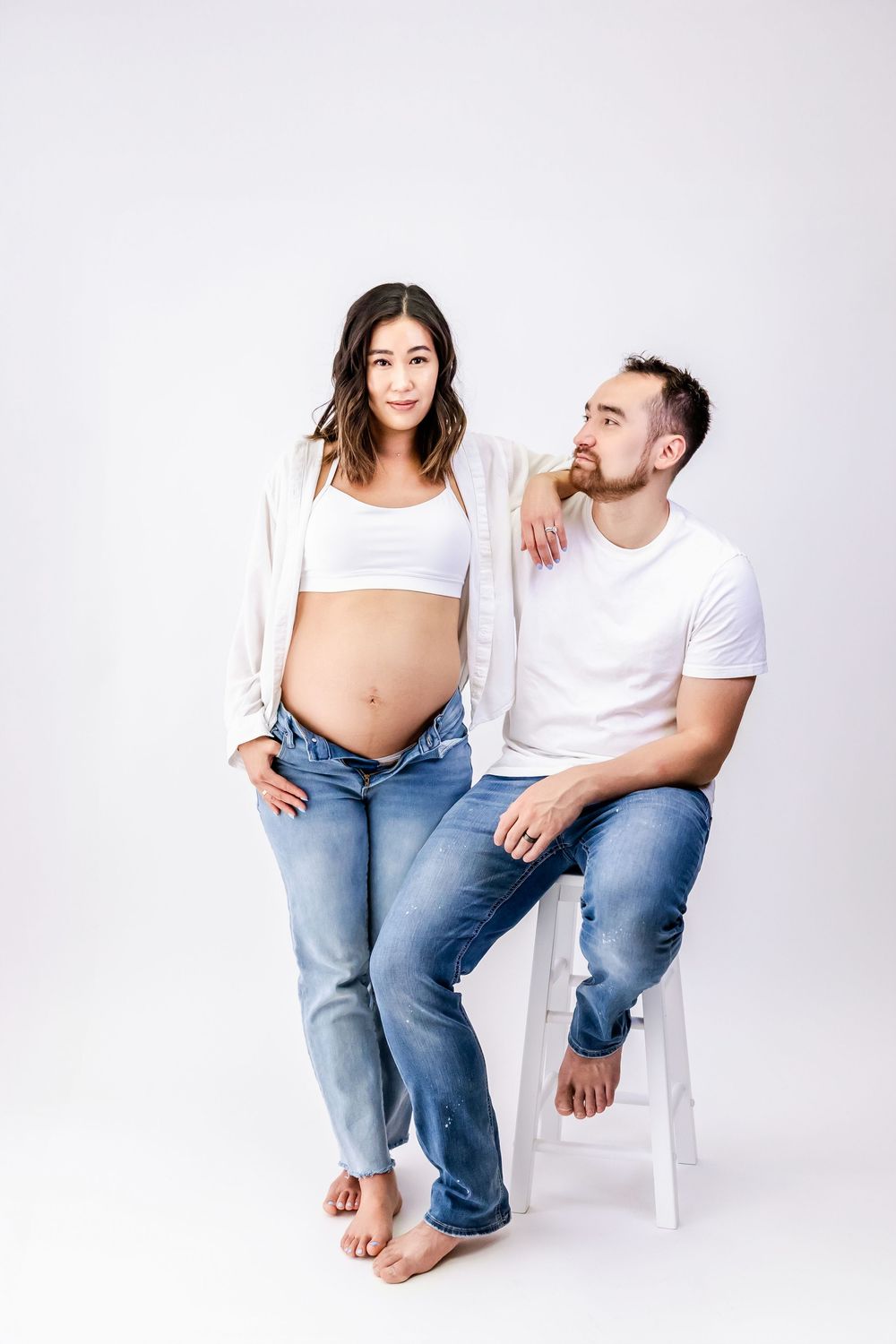 A couple wearing white tops and jeans pose casually barefoot on a white stool against a light background.
