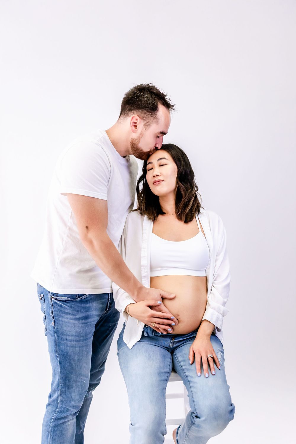 A couple in casual white tops and jeans share an intimate moment as they embrace against a light gray background.