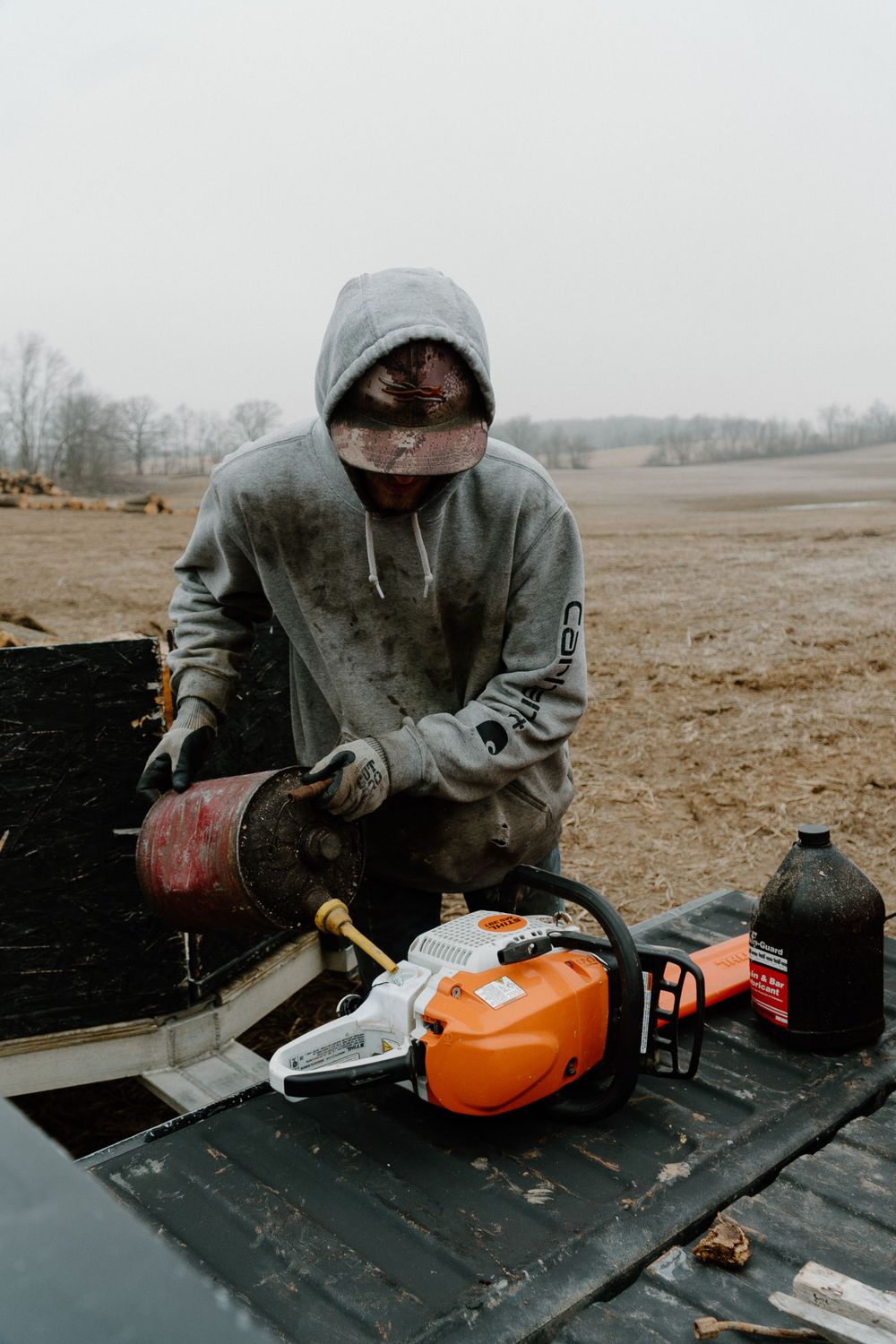 Worker in protective gear servicing chainsaw equipment on truck bed in muddy field.