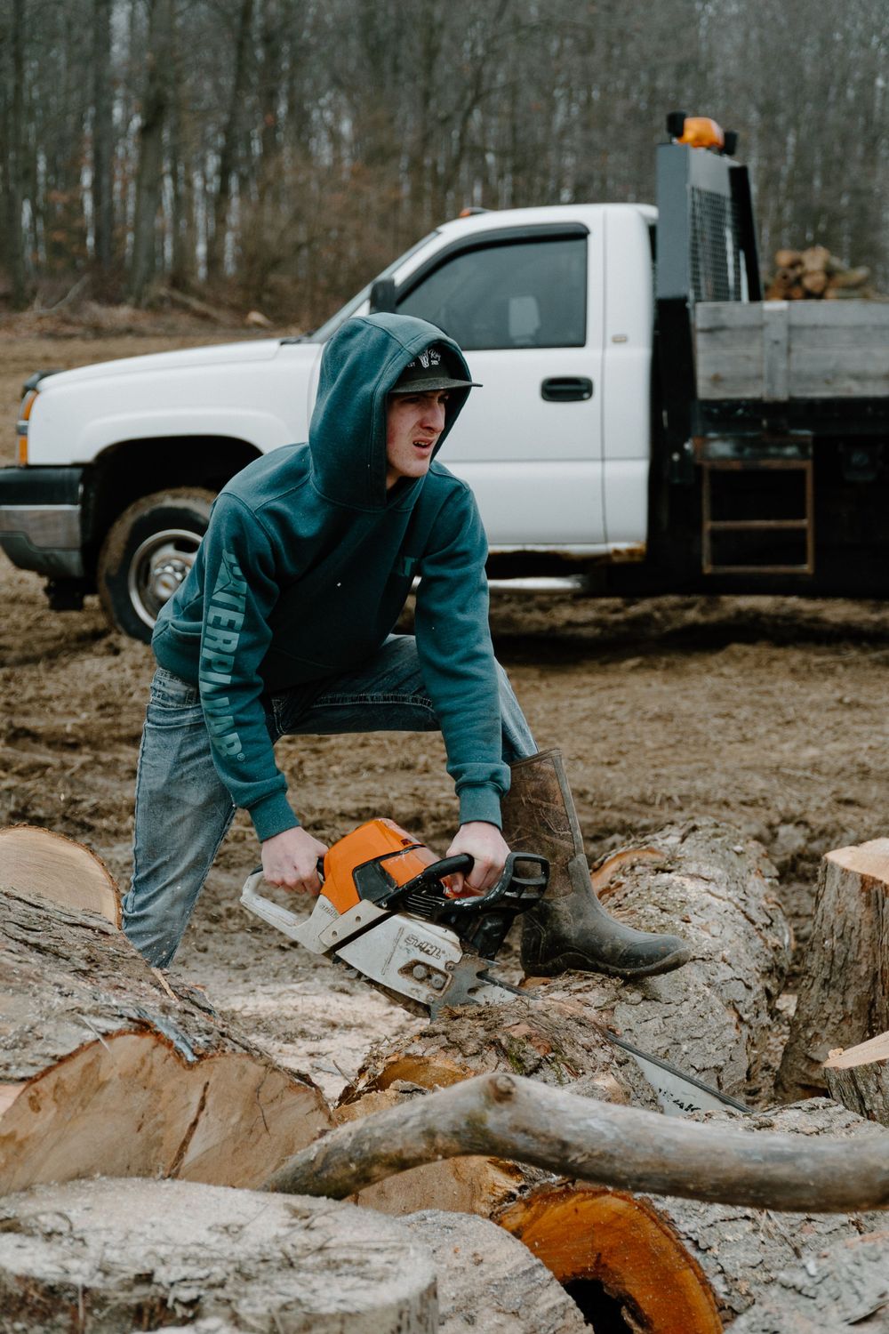 Person in teal hoodie operating chainsaw on cut logs with white truck in background.