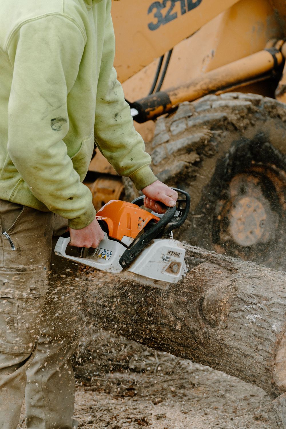 Person operating a chainsaw near construction equipment with muddy ground visible.