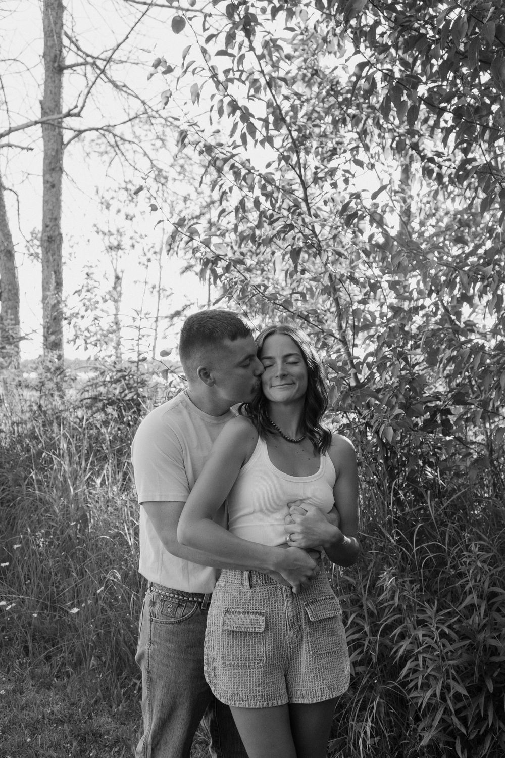Black and white portrait of couple sharing tender moment against leafy background.