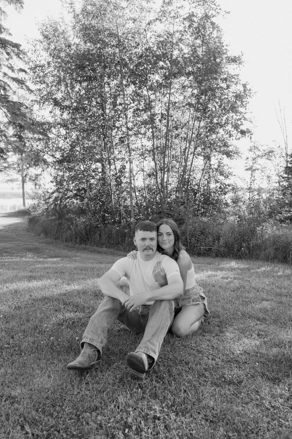 A couple sits together on grass with trees in the background in a black and white photo.