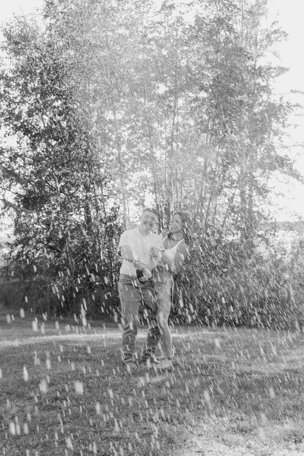 A couple plays in water droplets outdoors in a candid black and white moment.