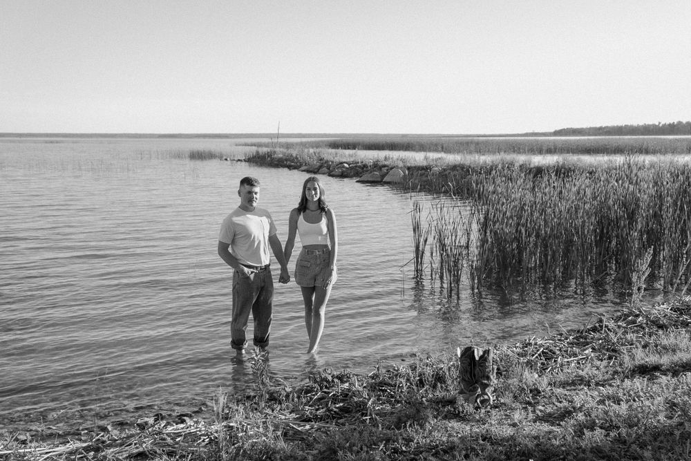 A couple walks together along the shoreline of a peaceful lake in black and white.