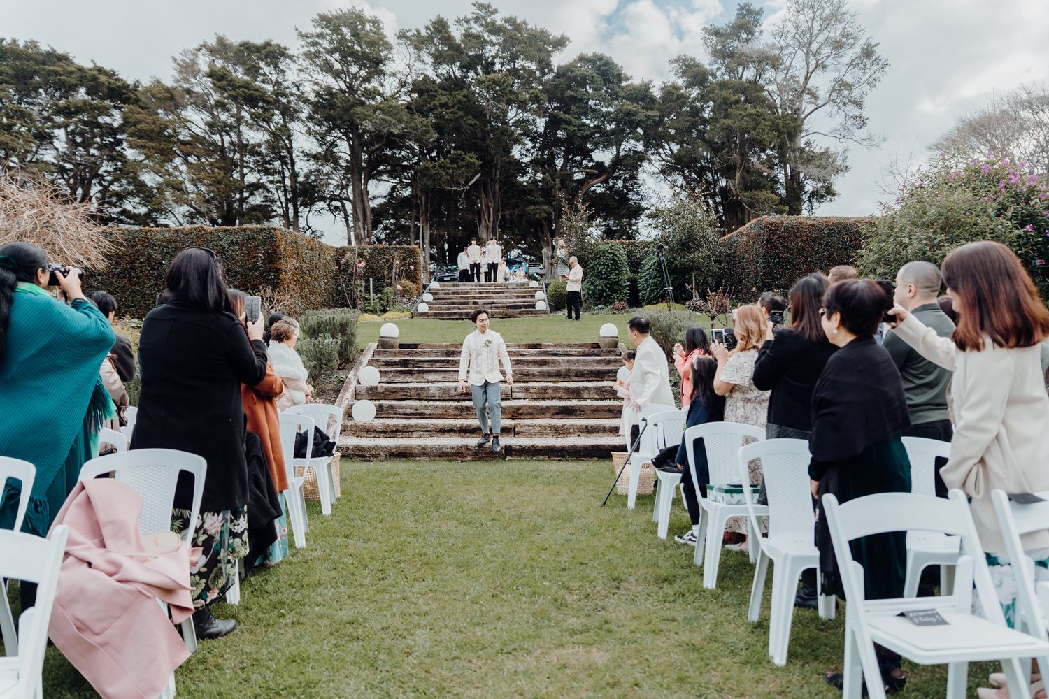 Wedding ceremony in progress with guests seated on white chairs on a grassy lawn with stone steps.