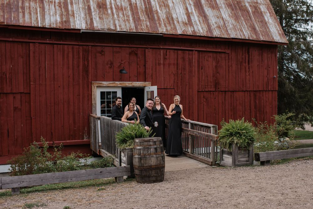 A rustic red barn with people gathered at the entrance alongside a wooden barrel and potted plants.