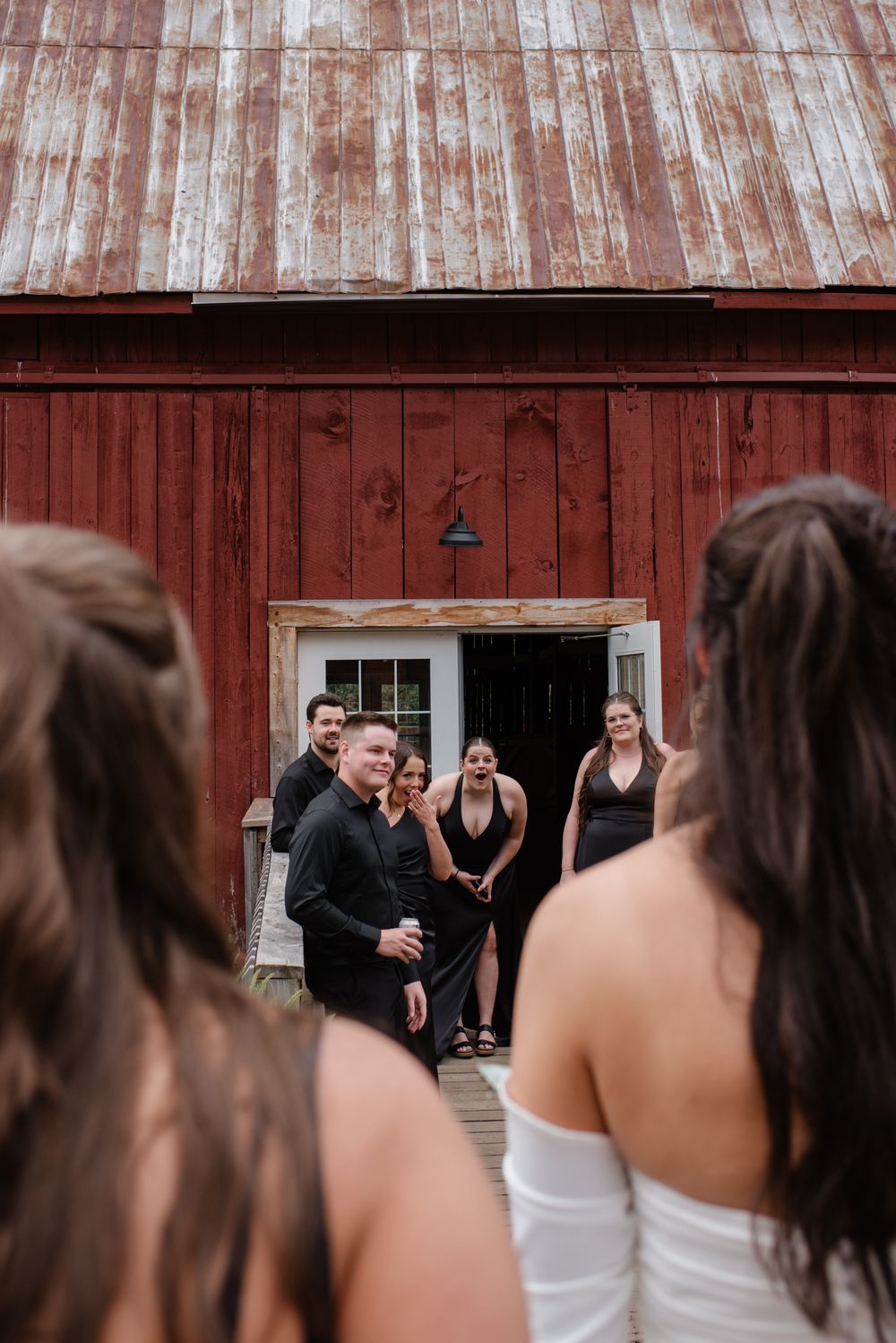A bridal party gathers outside a rustic red barn doorway during a wedding photoshoot.
