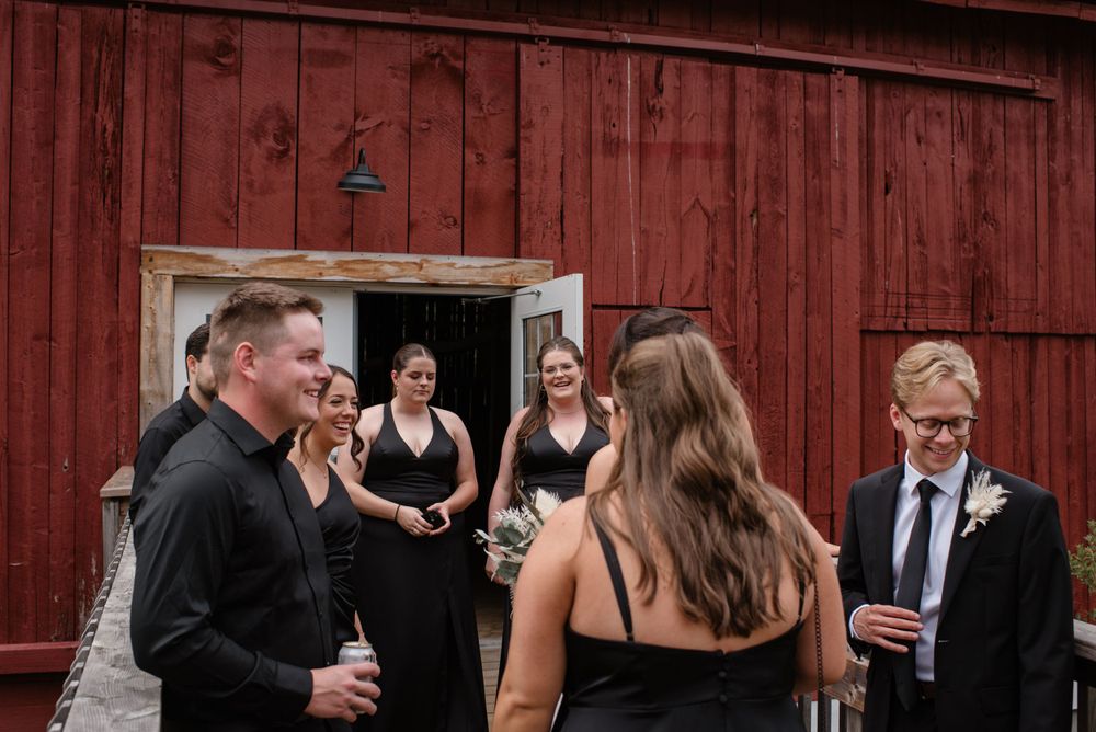 Wedding guests in formal attire gather and socialize outside a rustic red barn on a cloudy day.