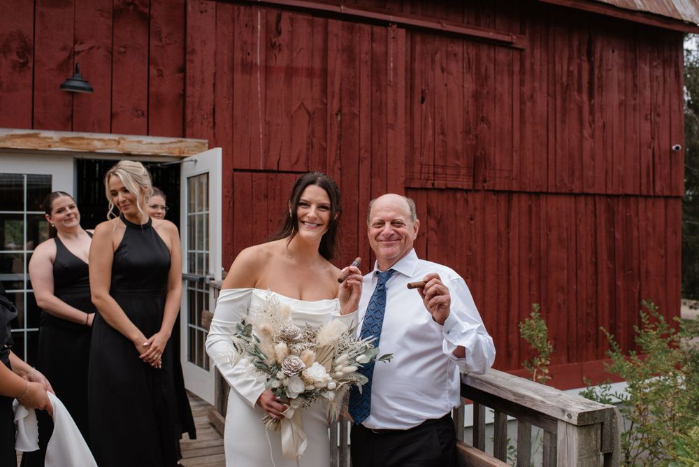 A group of formally dressed people pose in front of a rustic red barn on a wedding day.
