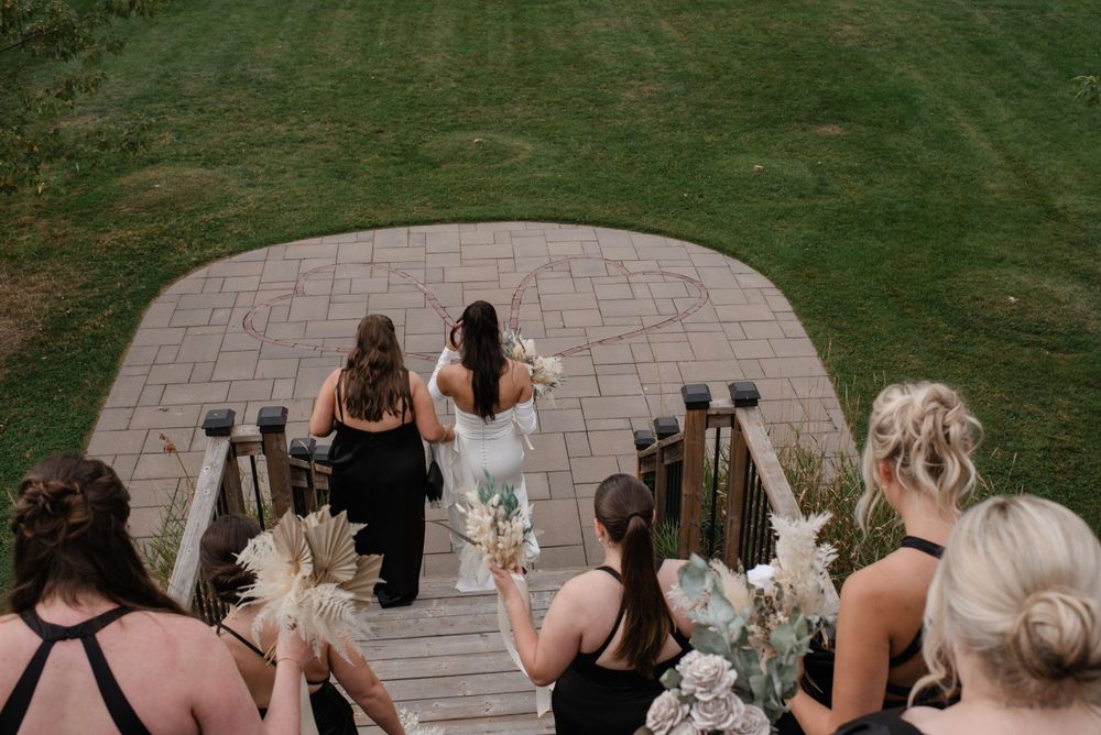 A bridal party descends outdoor wooden stairs carrying white floral bouquets in black dresses.