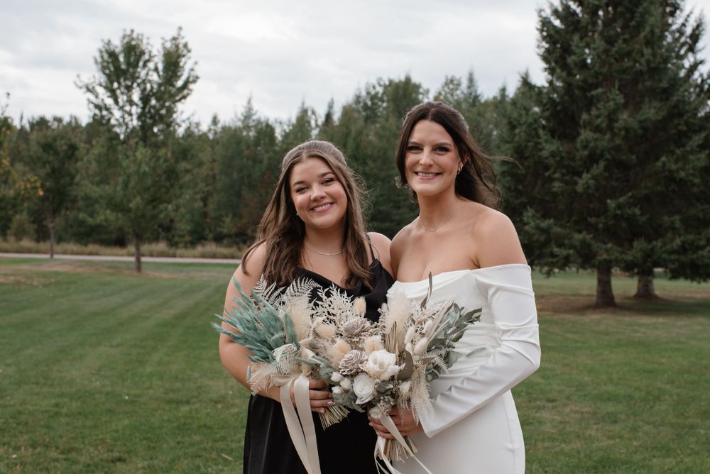 Two women pose outdoors holding delicate white and green bouquets against a backdrop of evergreen trees.