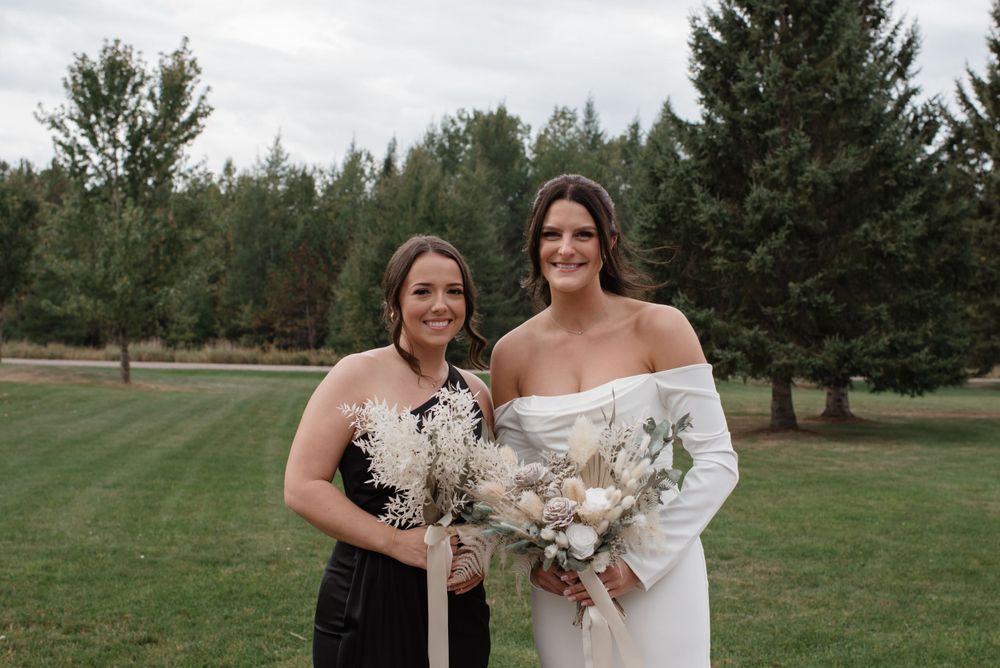 Two people in formal attire holding elegant floral bouquets pose together on a green lawn with evergreen trees in background.