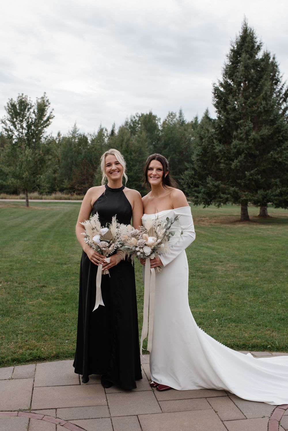 Two wedding attendants pose together on a garden path holding white floral bouquets against a backdrop of evergreen trees.