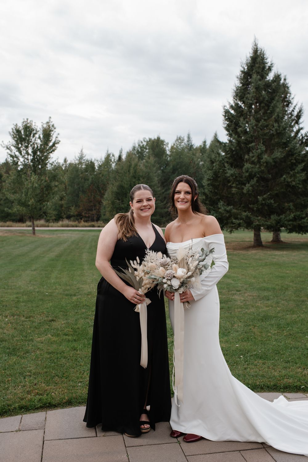 Two people pose outdoors in formal attire at a wedding with evergreen trees and grass in the background.