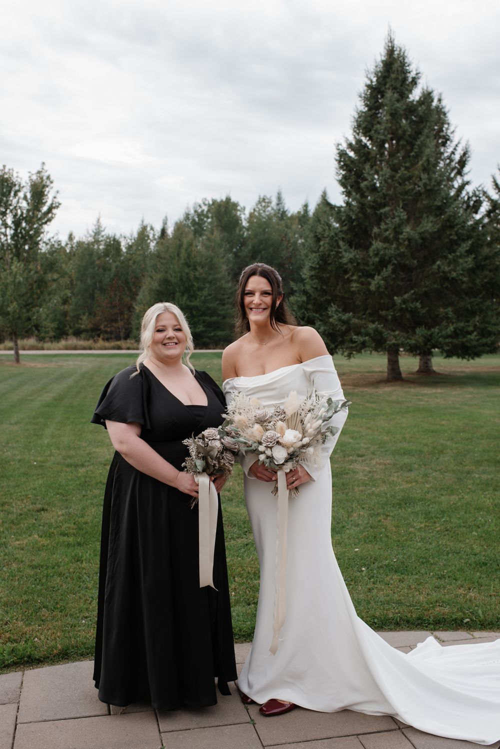 Two women in formal dresses, one in black and one in white, stand together holding bouquets on a lawn with evergreen trees.