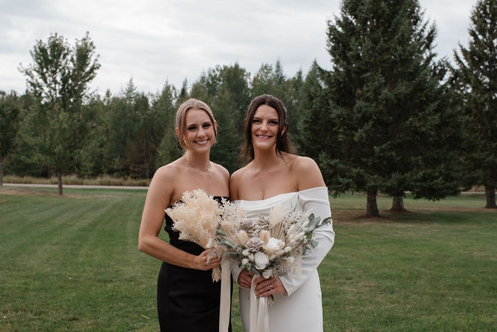 Two people in formal attire stand together holding white bouquets on a grassy lawn with evergreen trees in the background.