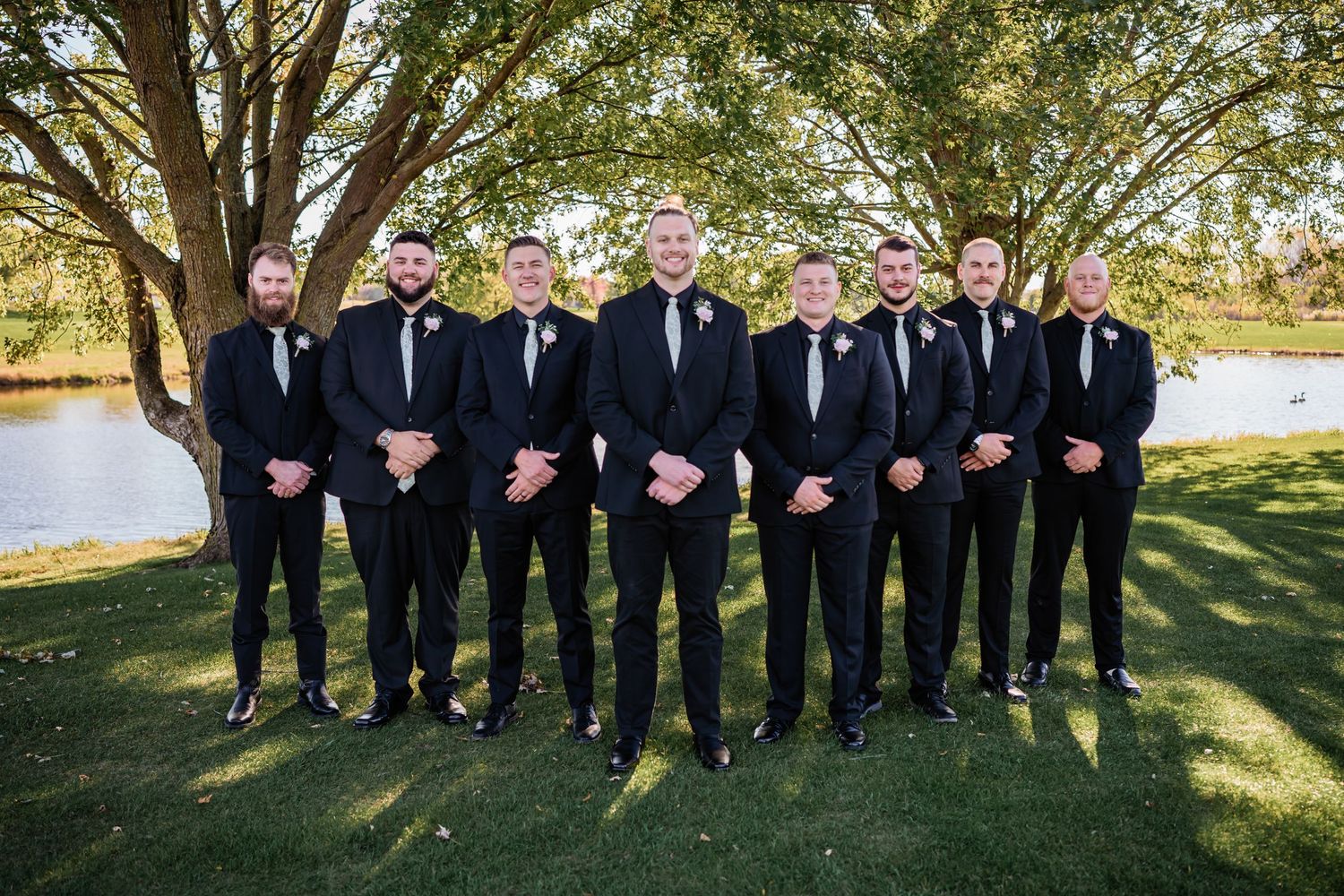 A wedding party of groomsmen in black suits and ties pose together on a sunny lawn beside a lake.