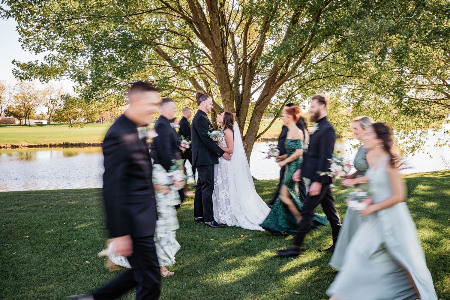 A wedding party gathers under a large tree beside a pond on a sunny afternoon.