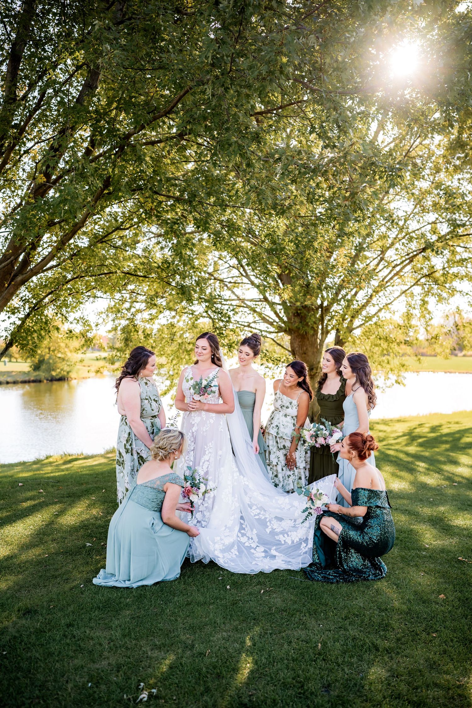 A wedding party gathered under sunlit trees by a lake wearing floral bridesmaid dresses and a white lace gown.