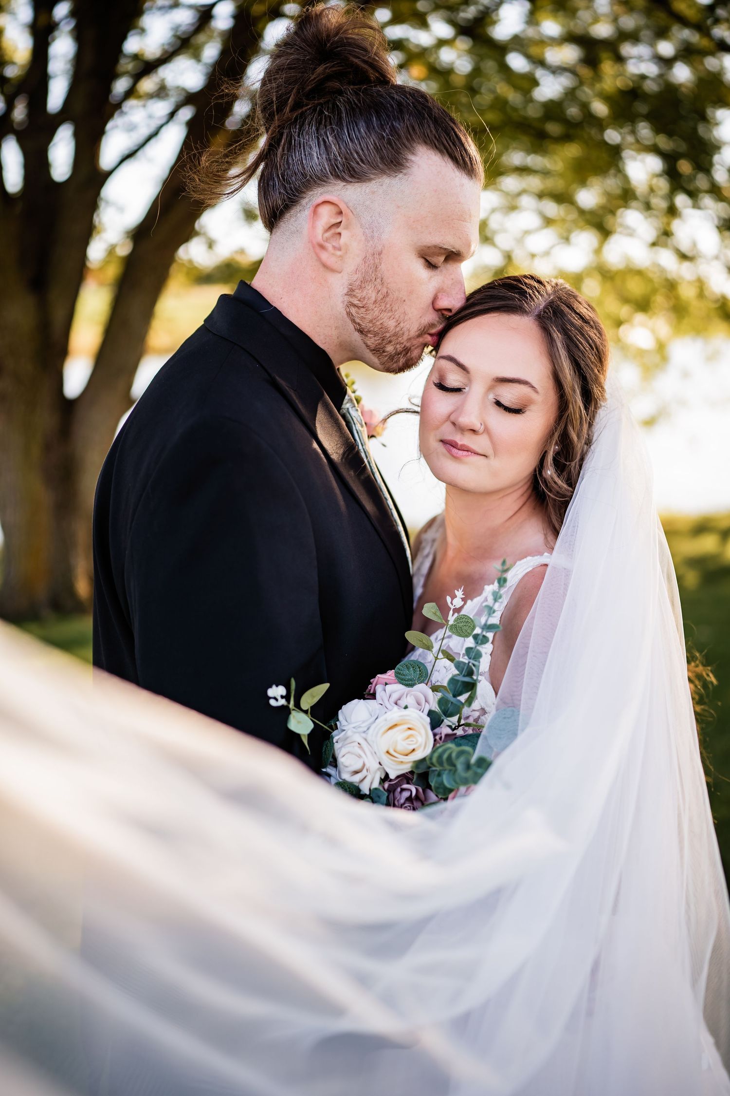 Newlywed couple shares intimate moment during outdoor wedding photography with flowing veil and pink floral bouquet.