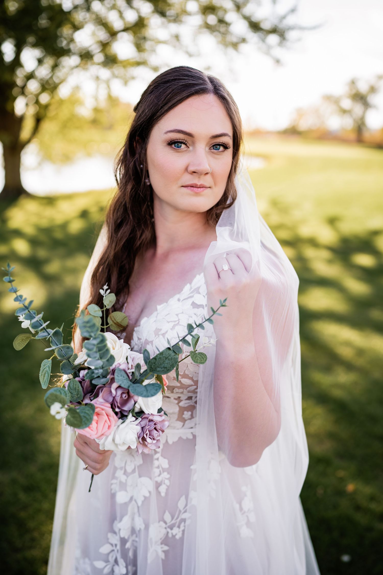 Beautiful bride in a delicate lace wedding gown holds soft pink and eucalyptus bouquet in outdoor garden setting.