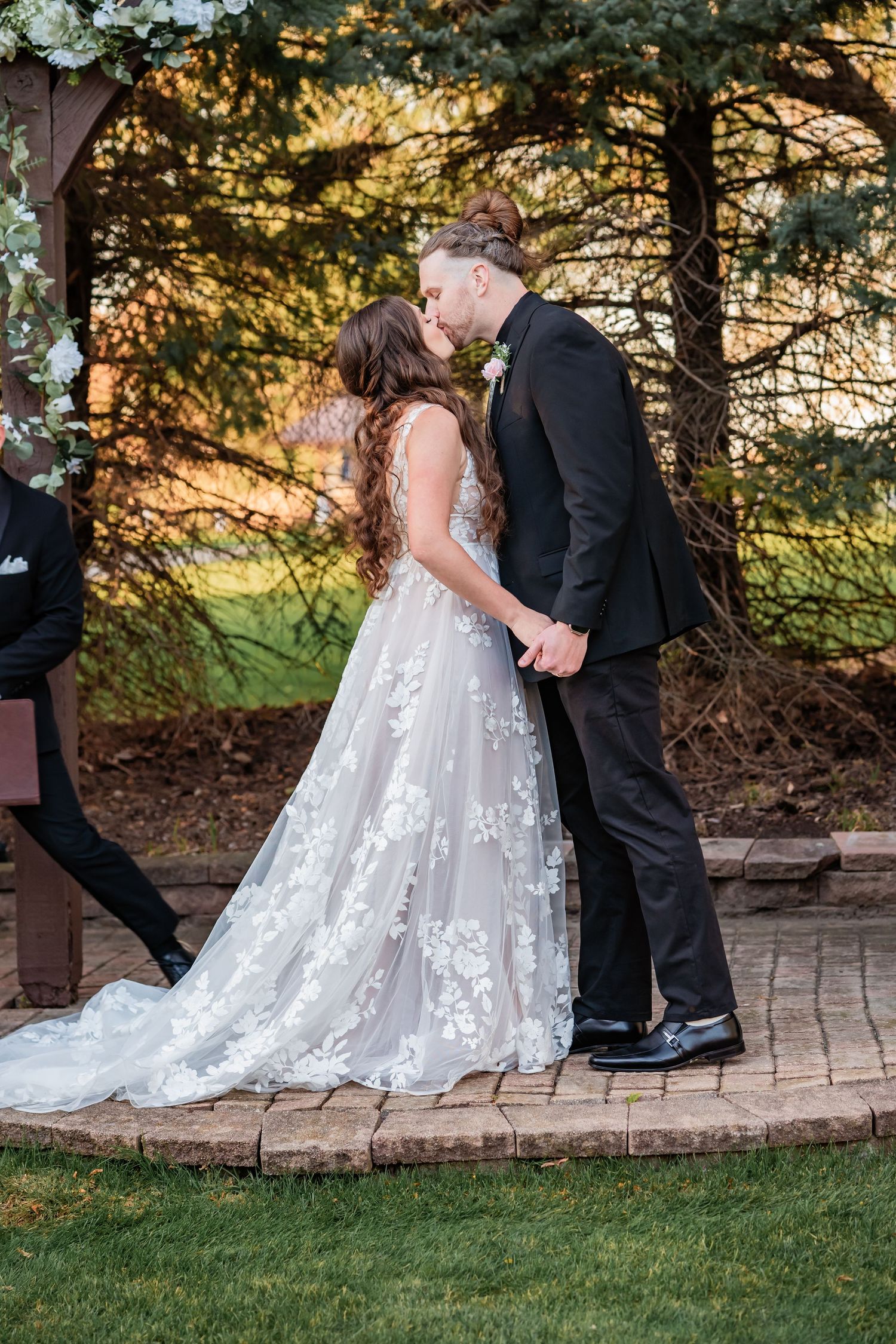 A couple shares their first kiss during a wedding ceremony on a brick pathway surrounded by evergreen trees at sunset.