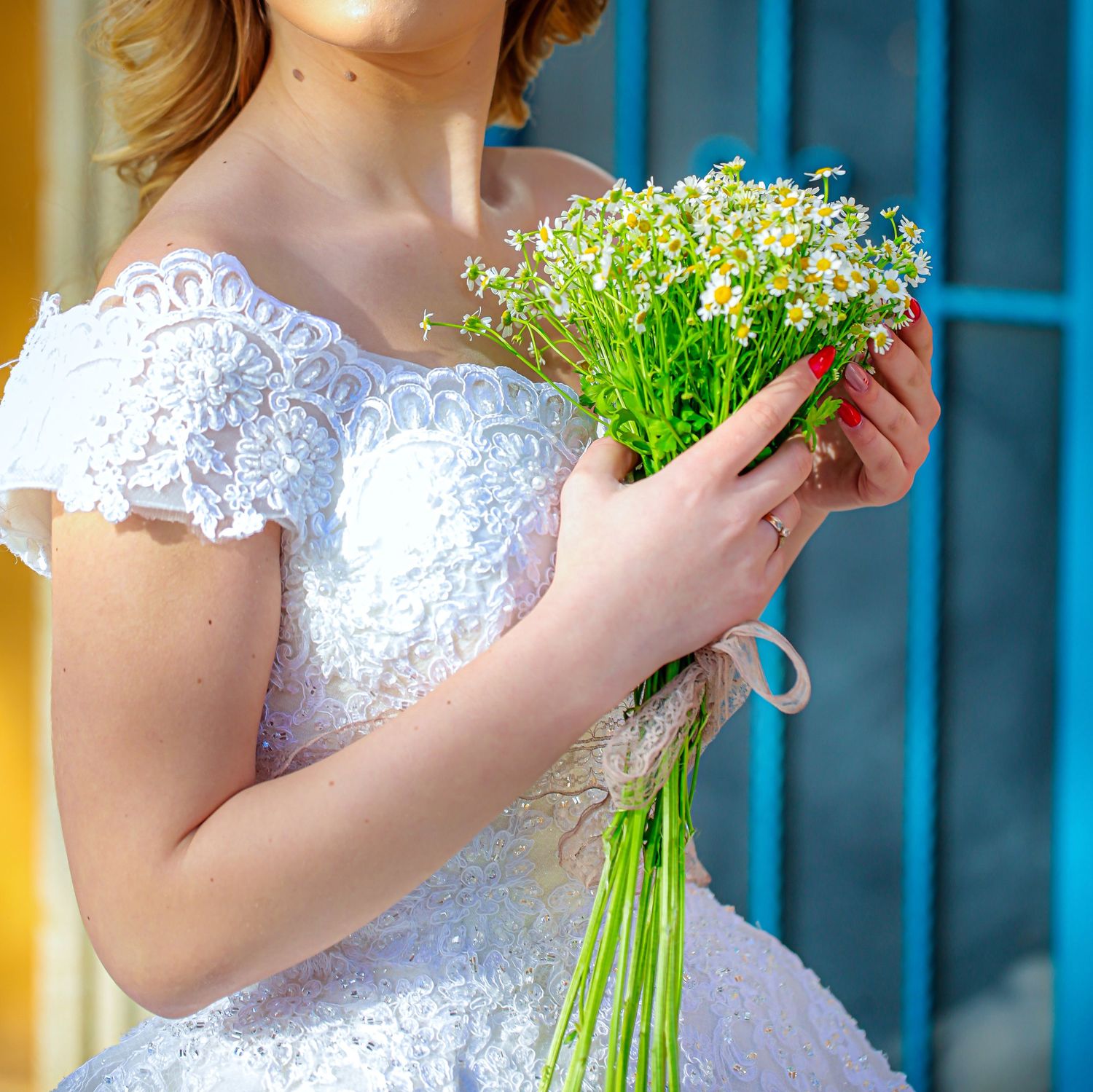 White lace wedding dress with delicate sleeves shown holding a fresh bouquet of small white wildflowers against a blue background.