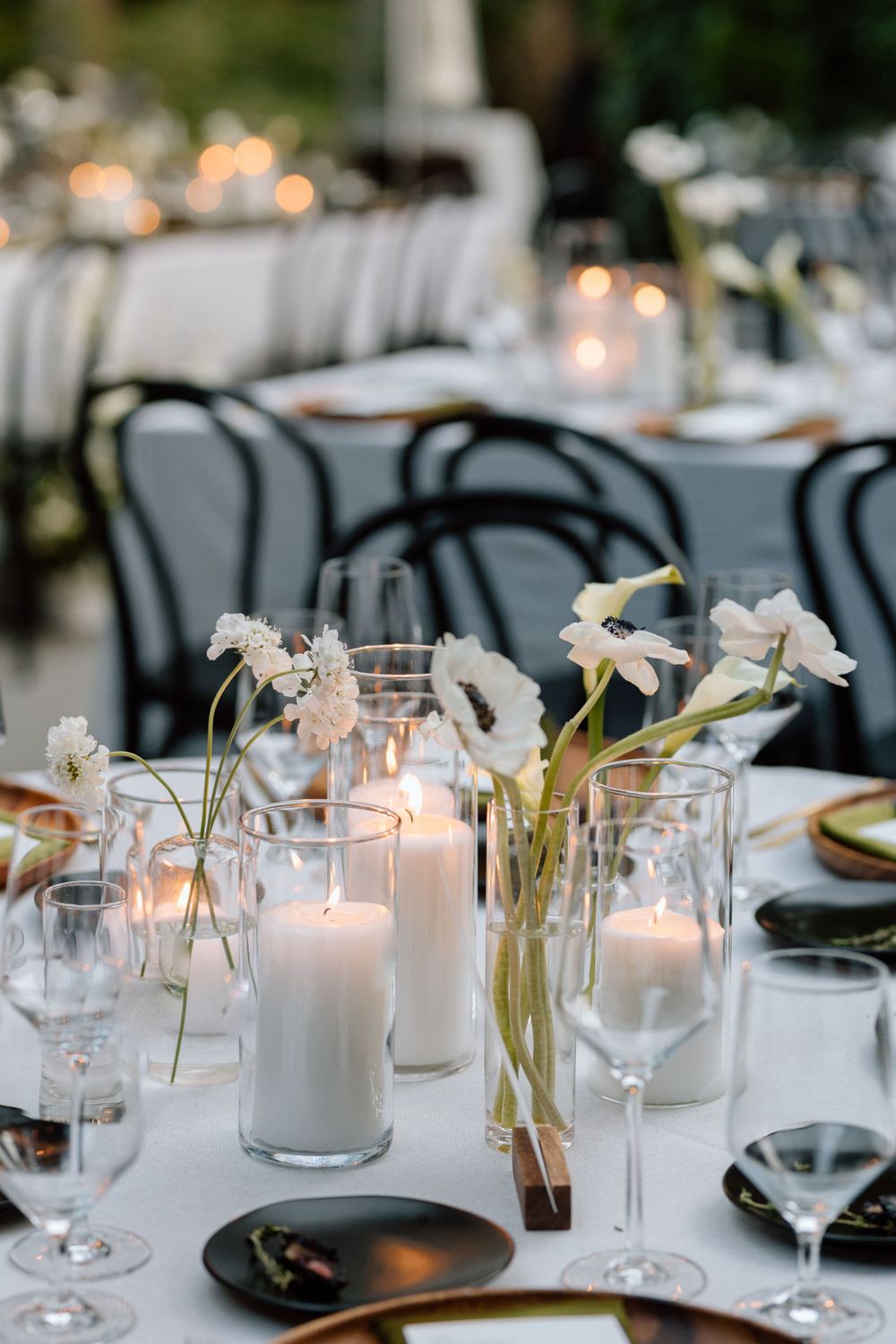 Elegant table setting with white candlesticks, floral arrangements in glass vases, and black chairs at an outdoor reception.
