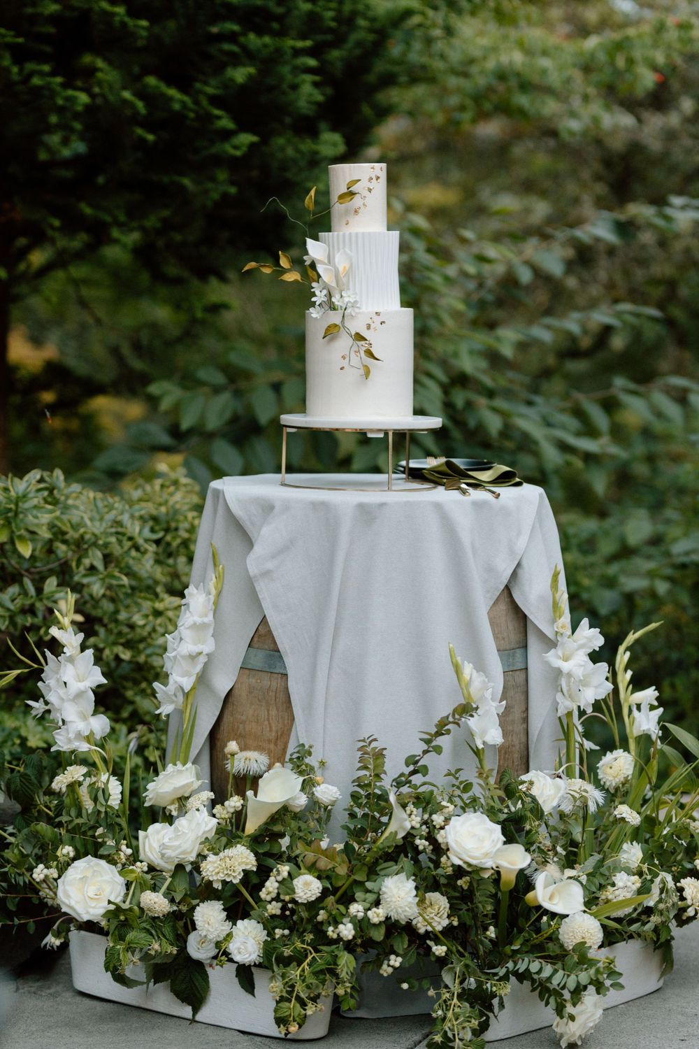 Elegant three-tier white wedding cake decorated with delicate greenery sits on a table surrounded by lush white floral arrangements.
