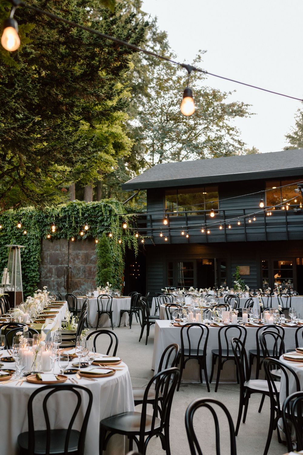 Elegant outdoor wedding reception setup with string lights, black bentwood chairs and white tablecloths on a patio.