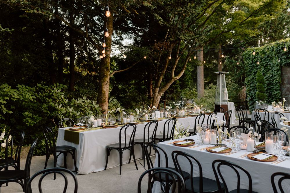 Elegant outdoor wedding reception setup with black bentwood chairs and white tablecloths under green garden foliage.