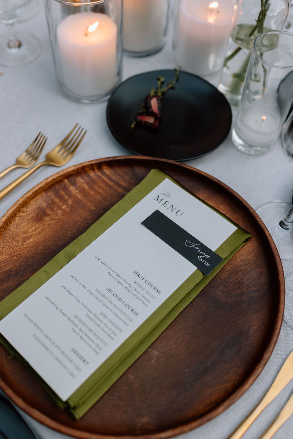 Elegant place setting with wooden charger plate, menu card with green accent, and glowing white candles on table.