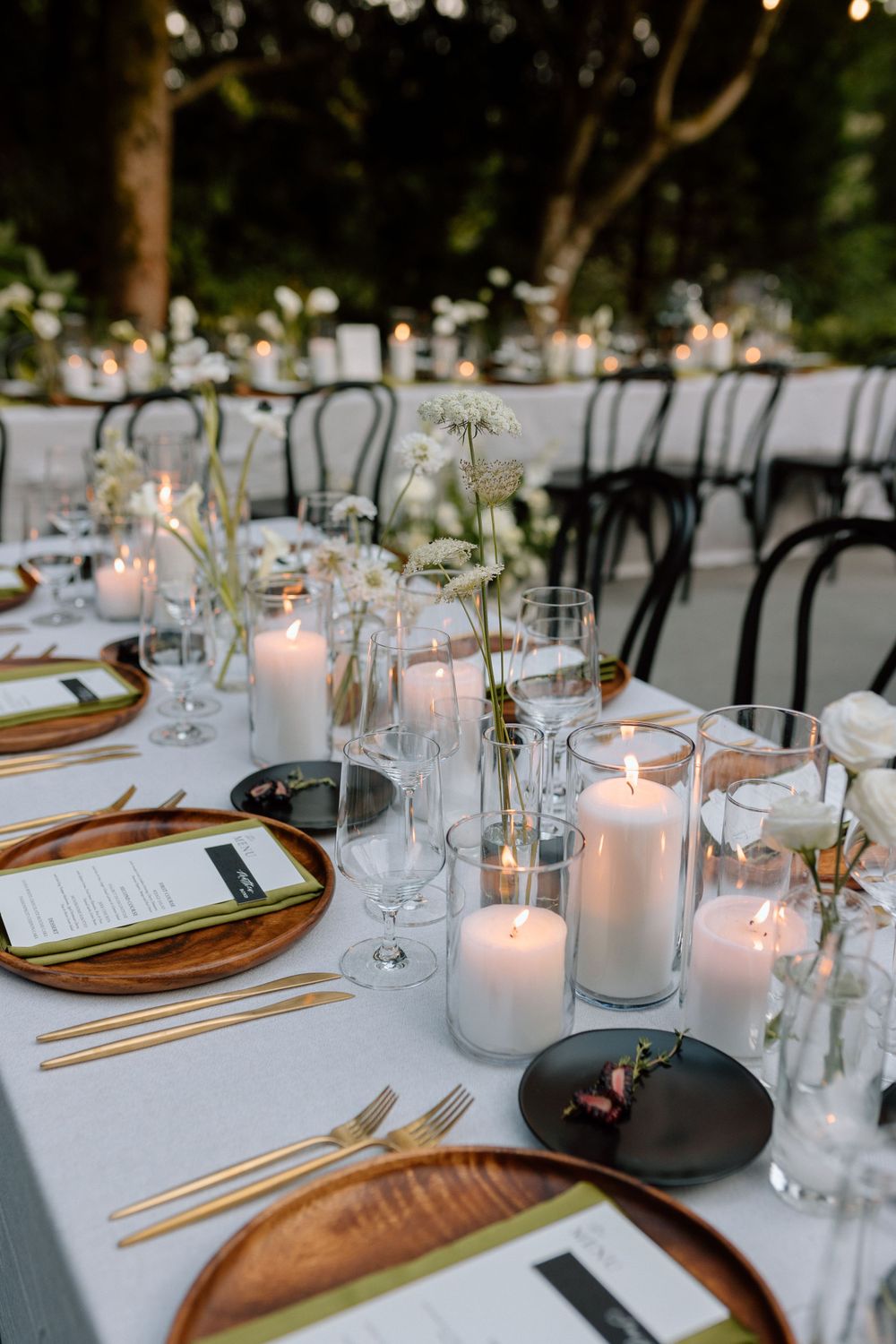Wedding reception table with modern place settings, white candles, and simple floral arrangements on outdoor patio.