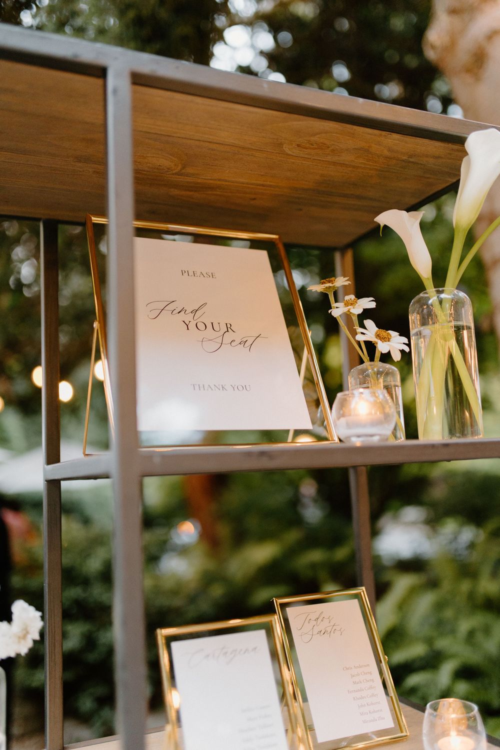 Elegant wedding reception decor with framed menus and white calla lilies displayed on wooden shelving with string lights.