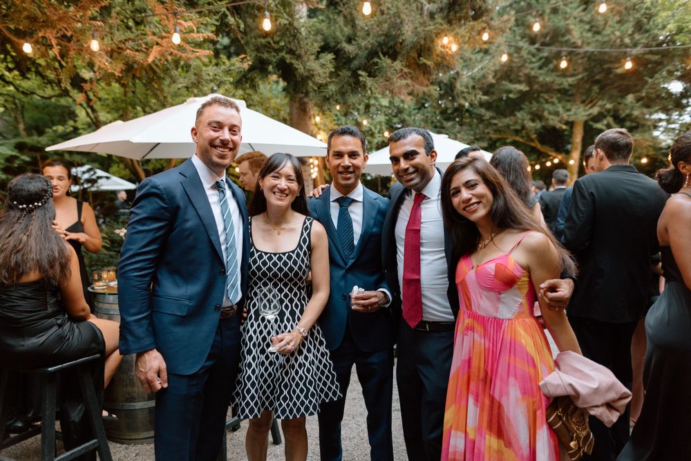 A group of well-dressed guests enjoying an outdoor wedding reception under trees with patio umbrellas.