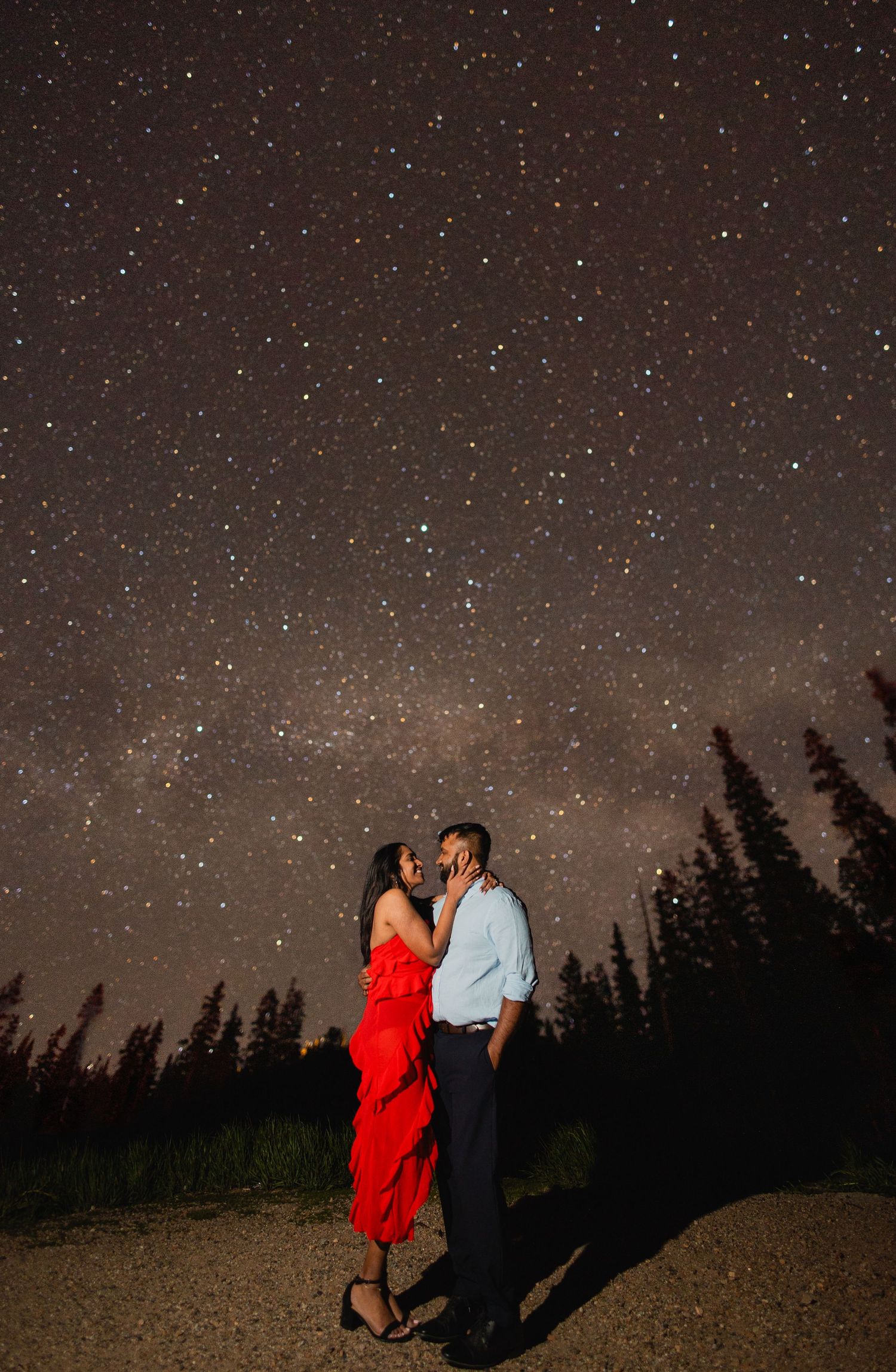 Two silhouetted figures embrace under a starlit sky with the Milky Way visible above pine trees at night.
