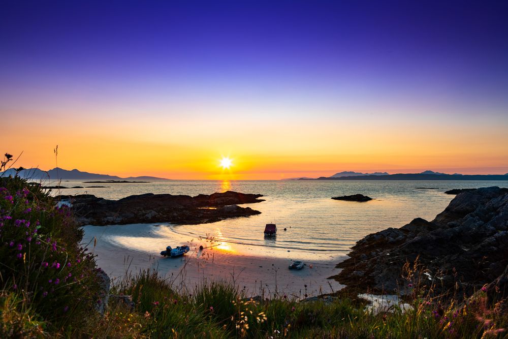 Stunning sunset over a coastal bay with rocky shores and calm waters reflecting golden light against purple sky.