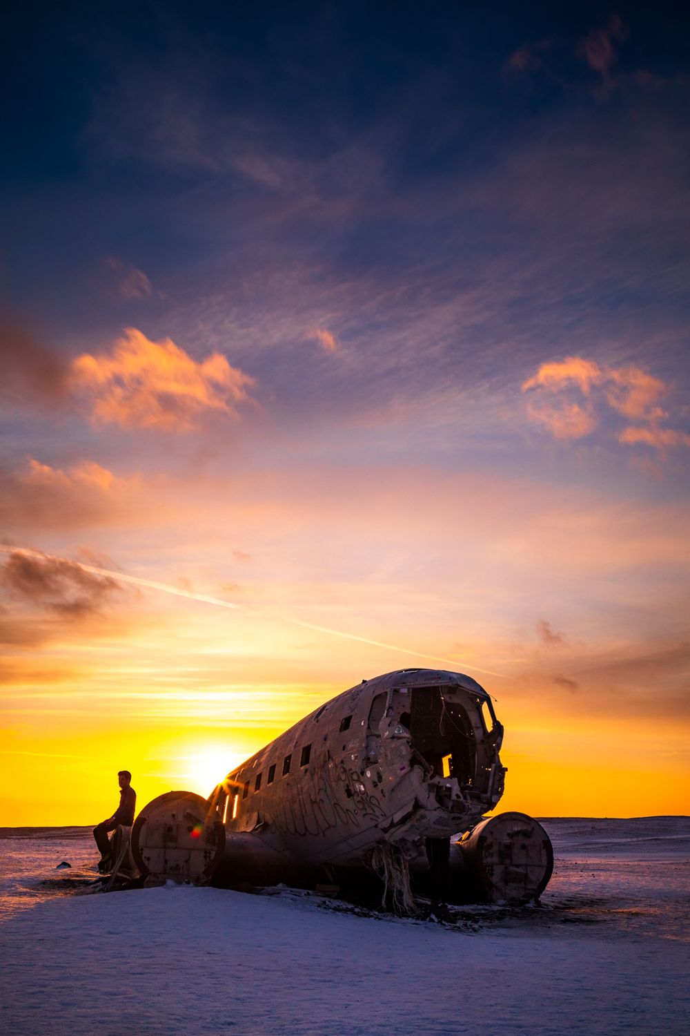 Abandoned airplane wreckage silhouetted against vibrant orange sunset sky on a snowy landscape in Iceland.