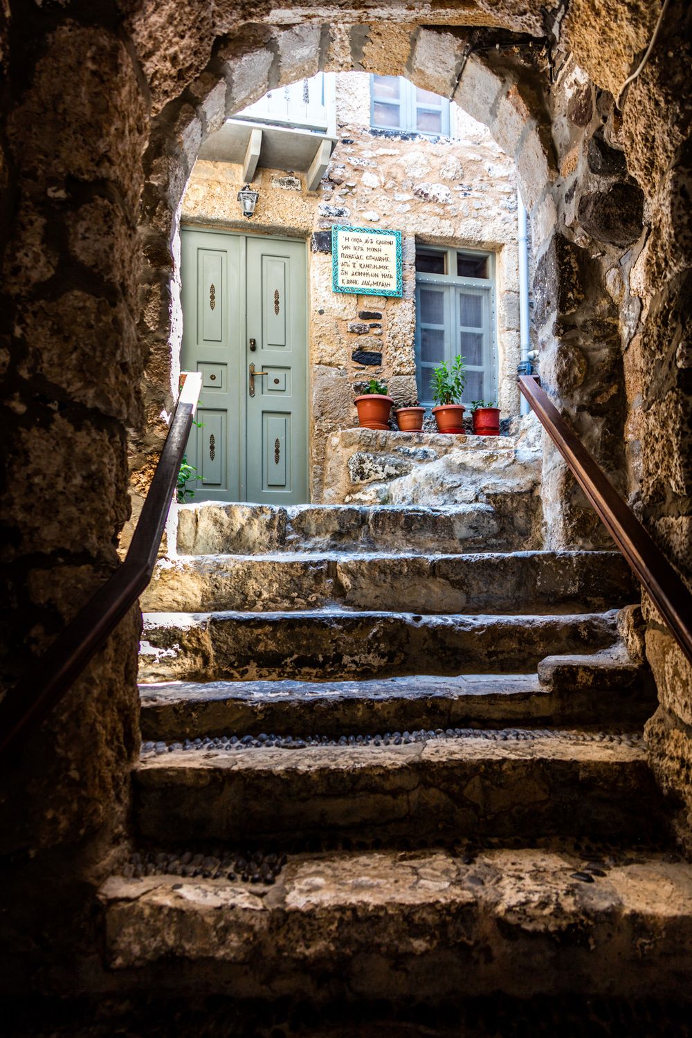 Stone archway frames rustic mint green door and window with terracotta pots on steps in Mediterranean village.