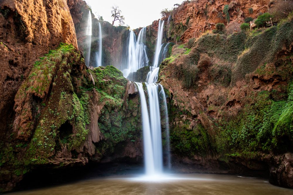 Scenic Ouzoud Falls cascade down moss-covered red cliffs into tranquil pool in Morocco's Atlas Mountains.