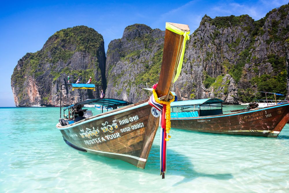 Traditional Thai long-tail boat anchored in crystal clear turquoise waters near limestone cliffs at Maya Bay, Thailand.