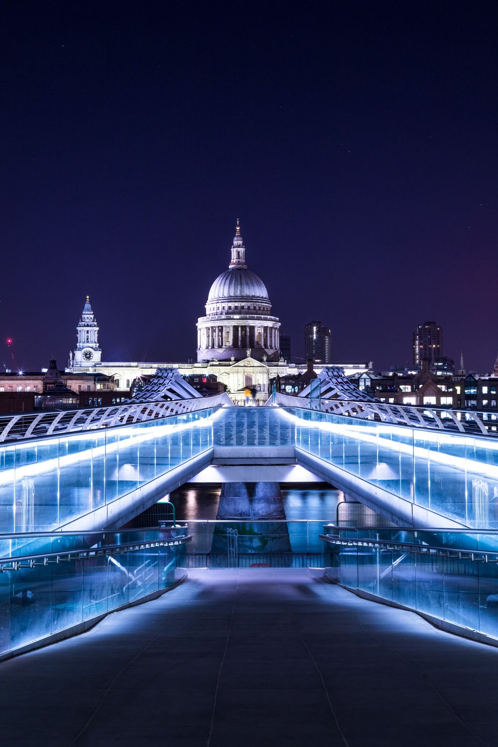 St Paul's Cathedral illuminated at night with Millennium Bridge lit in blue neon lights spanning the River Thames in London.