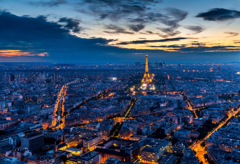 Aerial view of illuminated Paris cityscape at dusk with iconic Eiffel Tower glowing against dramatic sunset sky.