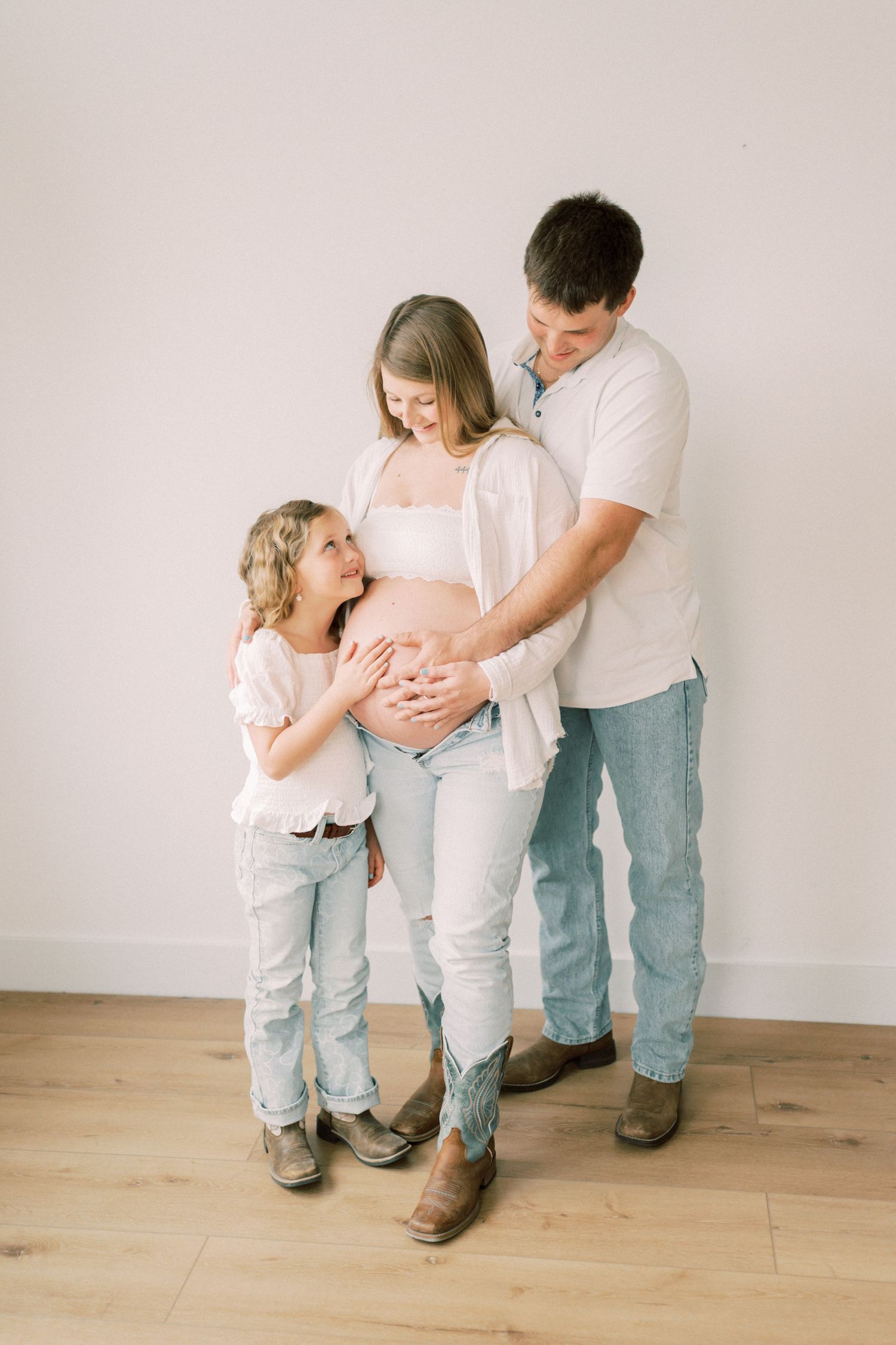 Family in matching white and denim outfits standing together and touching pregnant belly on wooden floor.
